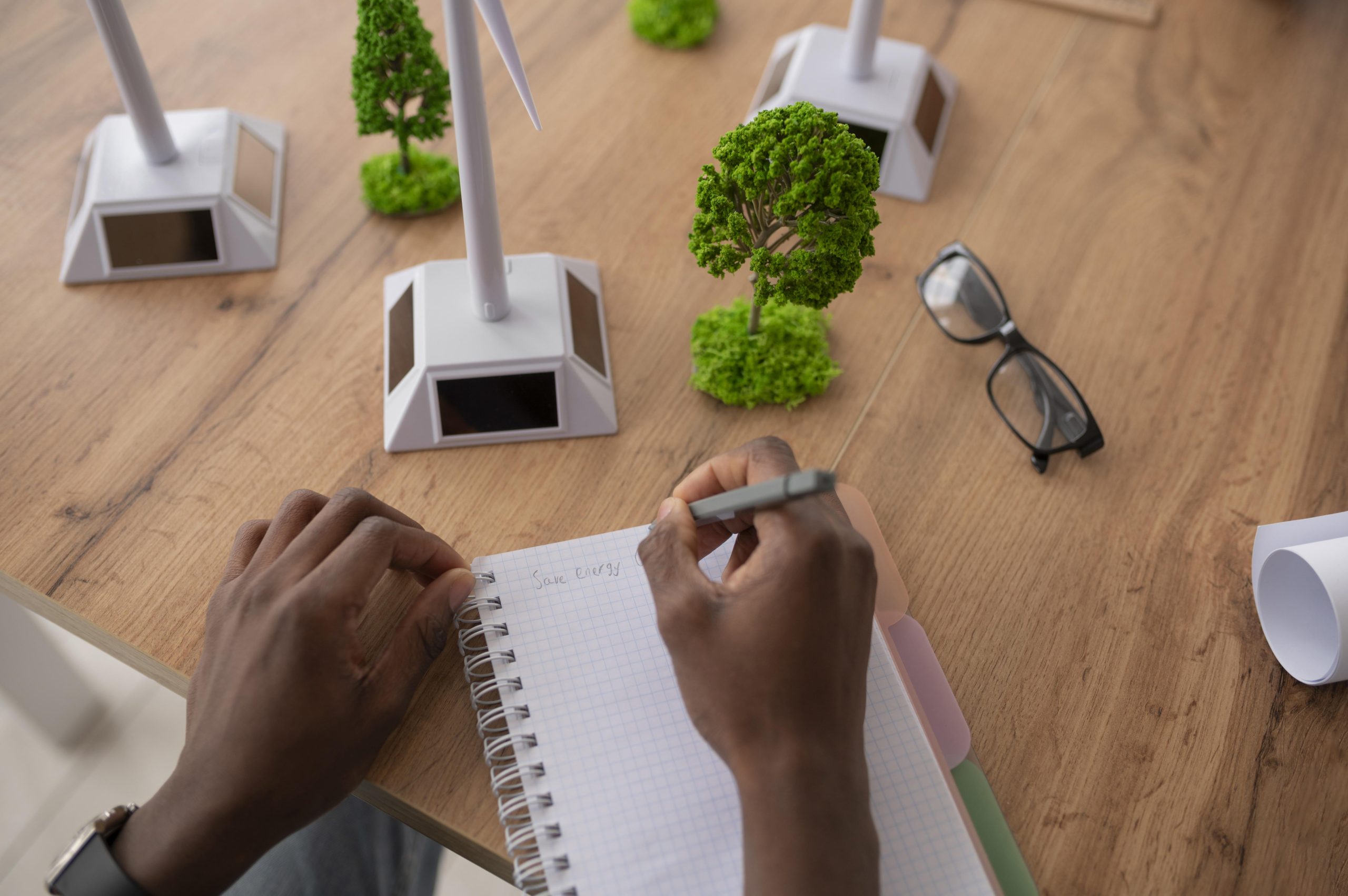 Mãos de um homem negro anotando em um caderno sobre uma mesa com maquetes de árvores e hélices eólicas