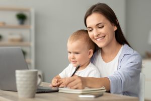 Estudar online: Jovem Mulher Segurando um Bebê e Estudando com Laptop em Casa.