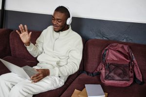 Estudante negro com fones de ouvido acenando para a câmera enquanto estuda online em um laptop sentado no sofá e interage socialmente com seus colegas.