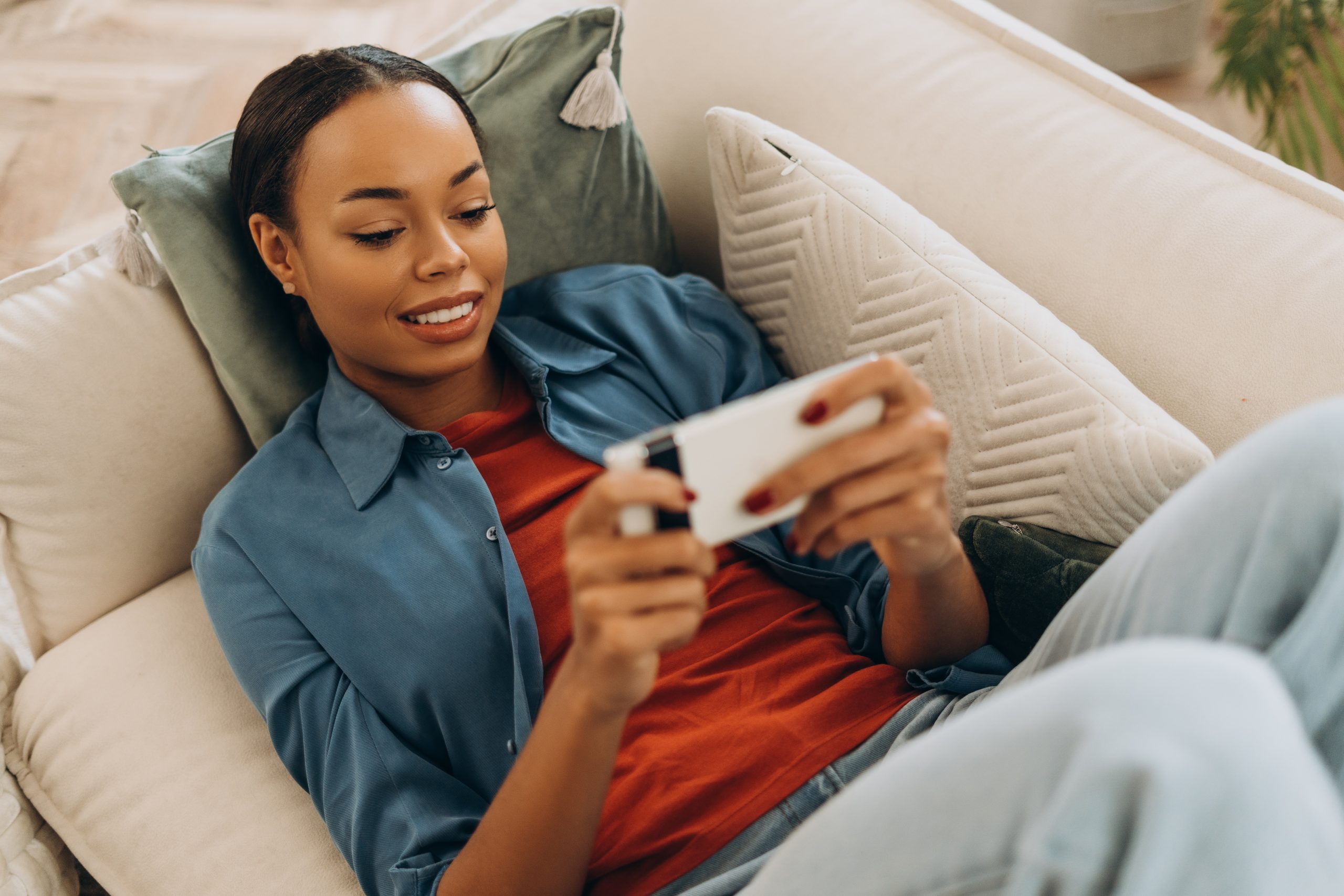 Uma jovem estudante negra assiste a uma aula no celular, deitada em seu sofá. Conceito: tecnologia na educação