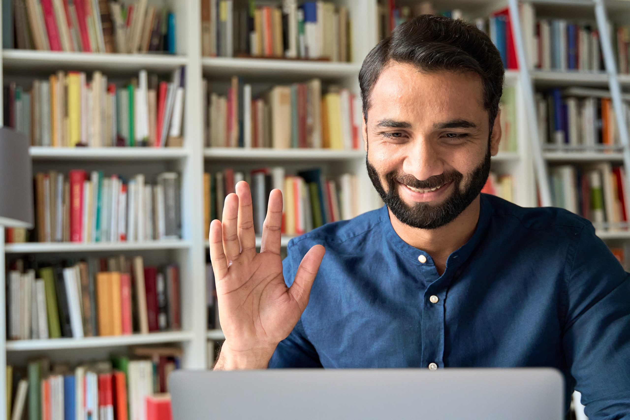 Homem com barba acenando com a mão para a câmera de seu laptop