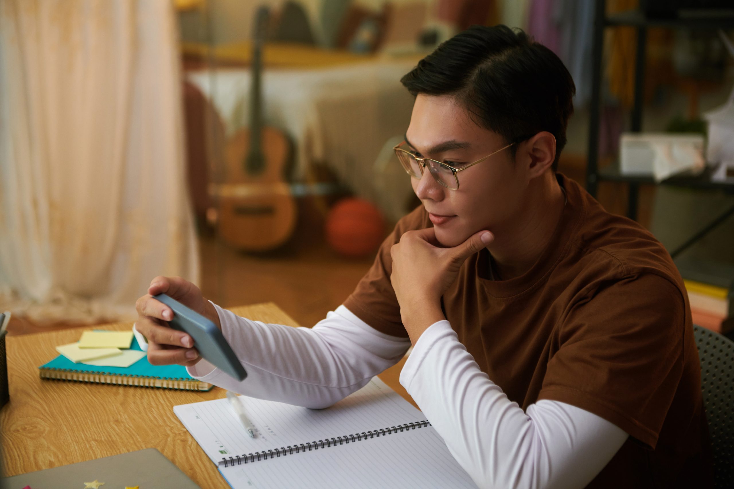 Um jovem asiático estuda pelo celular sentado à mesa, com um caderno aberto