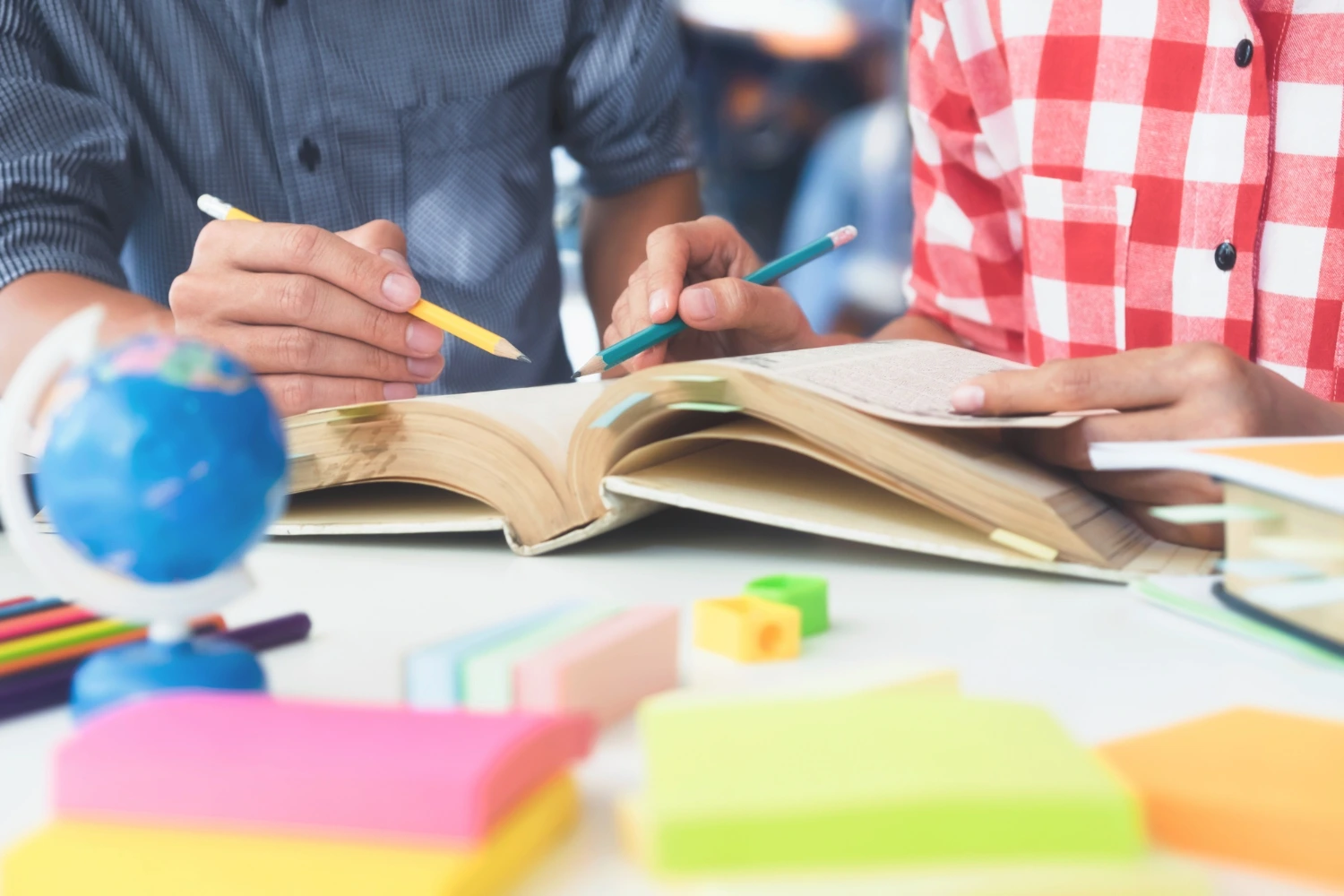 Duas pessoas estudando juntas com um livro aberto, ao lado de materiais escolares coloridos e um pequeno globo. Conceito: competências gerais da BNCC na prática.