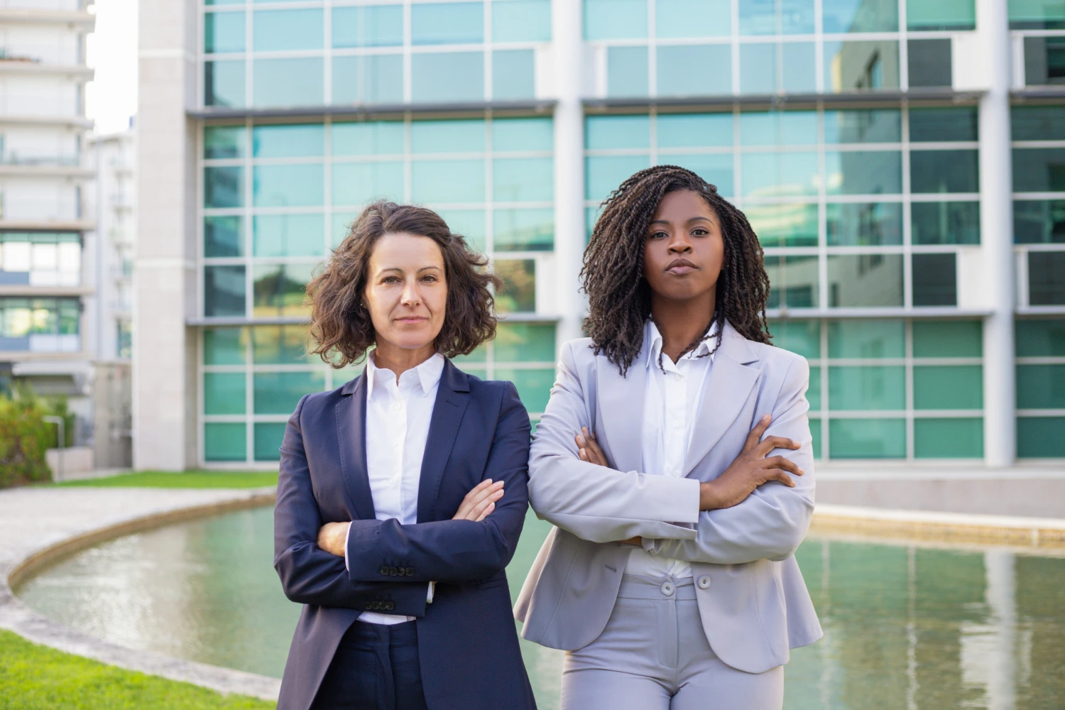 Duas mulheres executi vas de braços cruzadas em frente a uma empresa, representando equidade de gênero