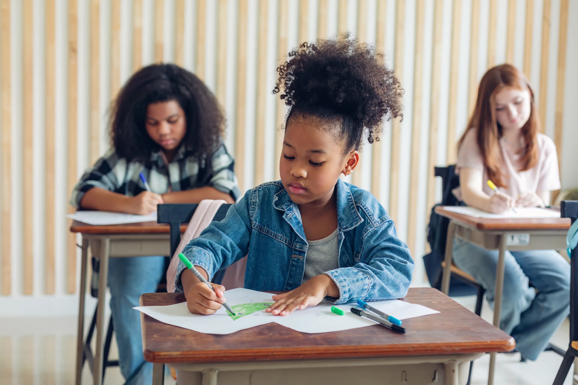 Crianças em uma sala de aula, focadas em atividades escolares, com destaque para uma menina desenhando. 