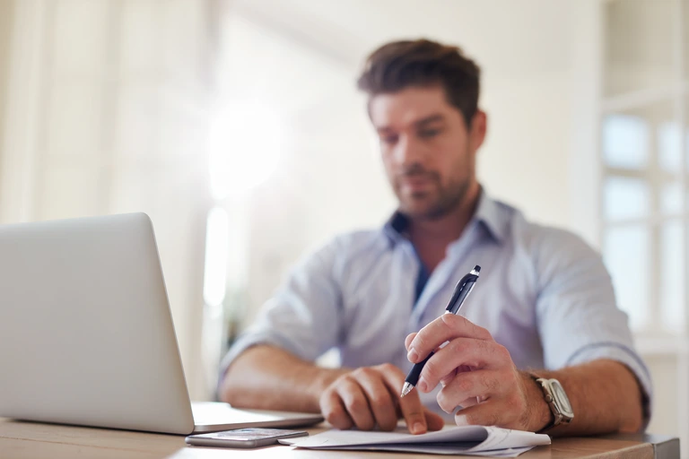 Um homem em ambiente de trabalho com o laptop aberto e fazendo anotações. Conceito: tipos de feedback - autofeedback