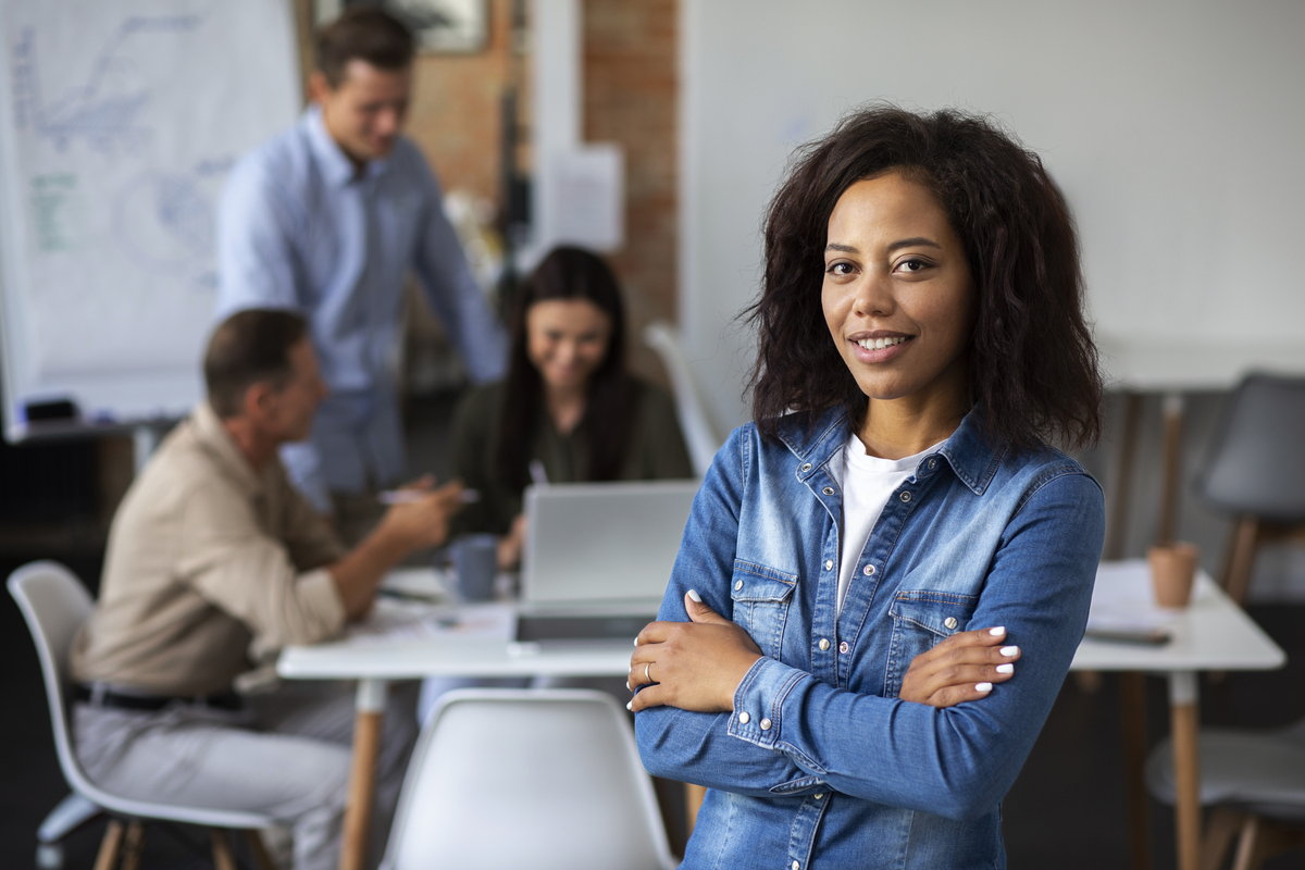 Uma mulher negra de braços cruzados e sorridente em uma sala de reuniões com sua equipe ao fundo.