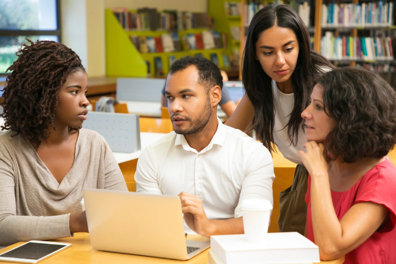 Quatro professores reunidos em biblioteca, em torno de um laptop, debatendo pontos de Projeto Político-Pedagógico