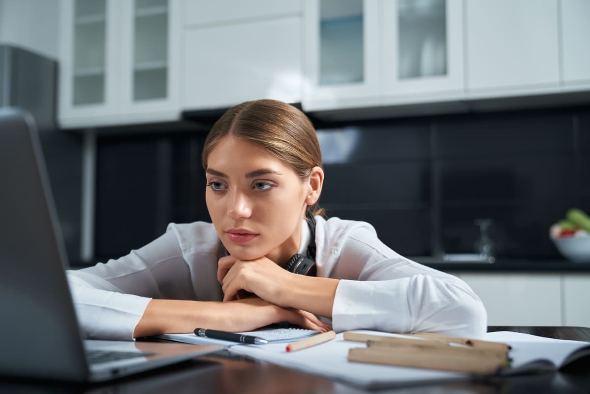 Mulher jovem com expressão pensativa apoia o rosto nas mãos enquanto observa a tela de um laptop. Sobre a mesa, cadernos, canetas e fones de ouvido indicam um momento de estudo ou trabalho remoto. O ambiente moderno da cozinha ao fundo sugere um espaço de home office. Representação do desenvolvimento de hard skills, como análise de dados, pesquisa e aprendizado online.