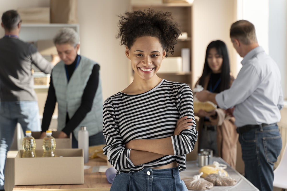 Mulher jovem sorridente de braços cruzados em primeiro plano, vestindo blusa listrada preta e branca. Ao fundo, um grupo de pessoas trabalha organizando doações, empacotando alimentos e suprimentos em uma sala iluminada. Representação de colaboração e trabalho voluntário, onde habilidades técnicas e organizacionais (hard skills) são aplicadas na logística da ação.