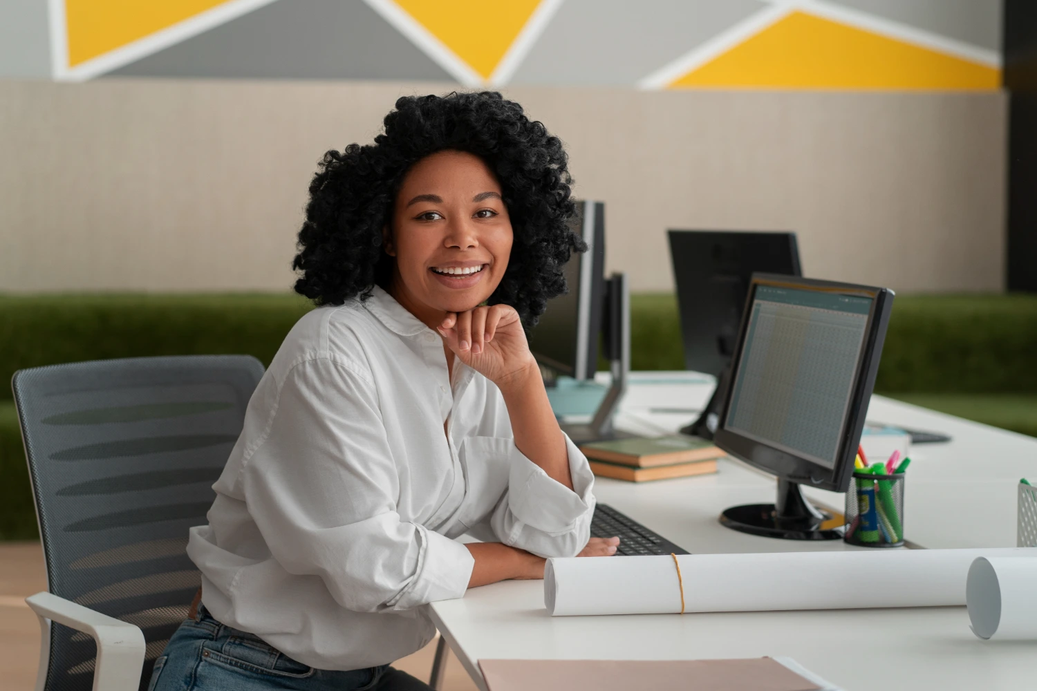 Mulher sorrindo em sua estação de trabalho com computadores e gráficos na tela, representando o trabalho digital de um assistente administrativo moderno.