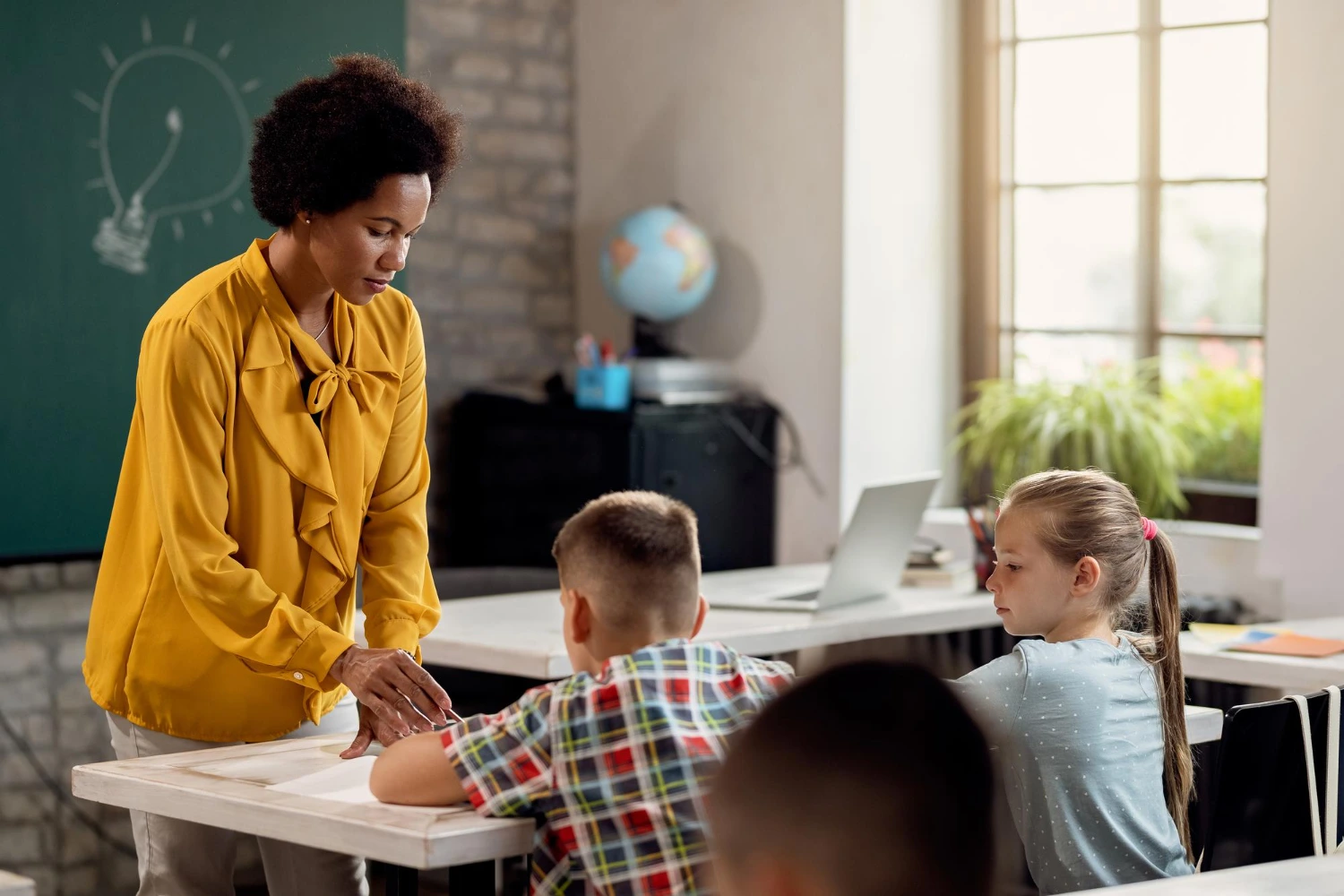 Uma professora vestida de amarelo está interagindo com alunos em uma sala de aula bem iluminada. Ela demonstra empatia e suporte a um aluno. A cena destaca a importância das habilidades emocionais na educação, como comunicação ativa e compreensão dos sentimentos dos outros.