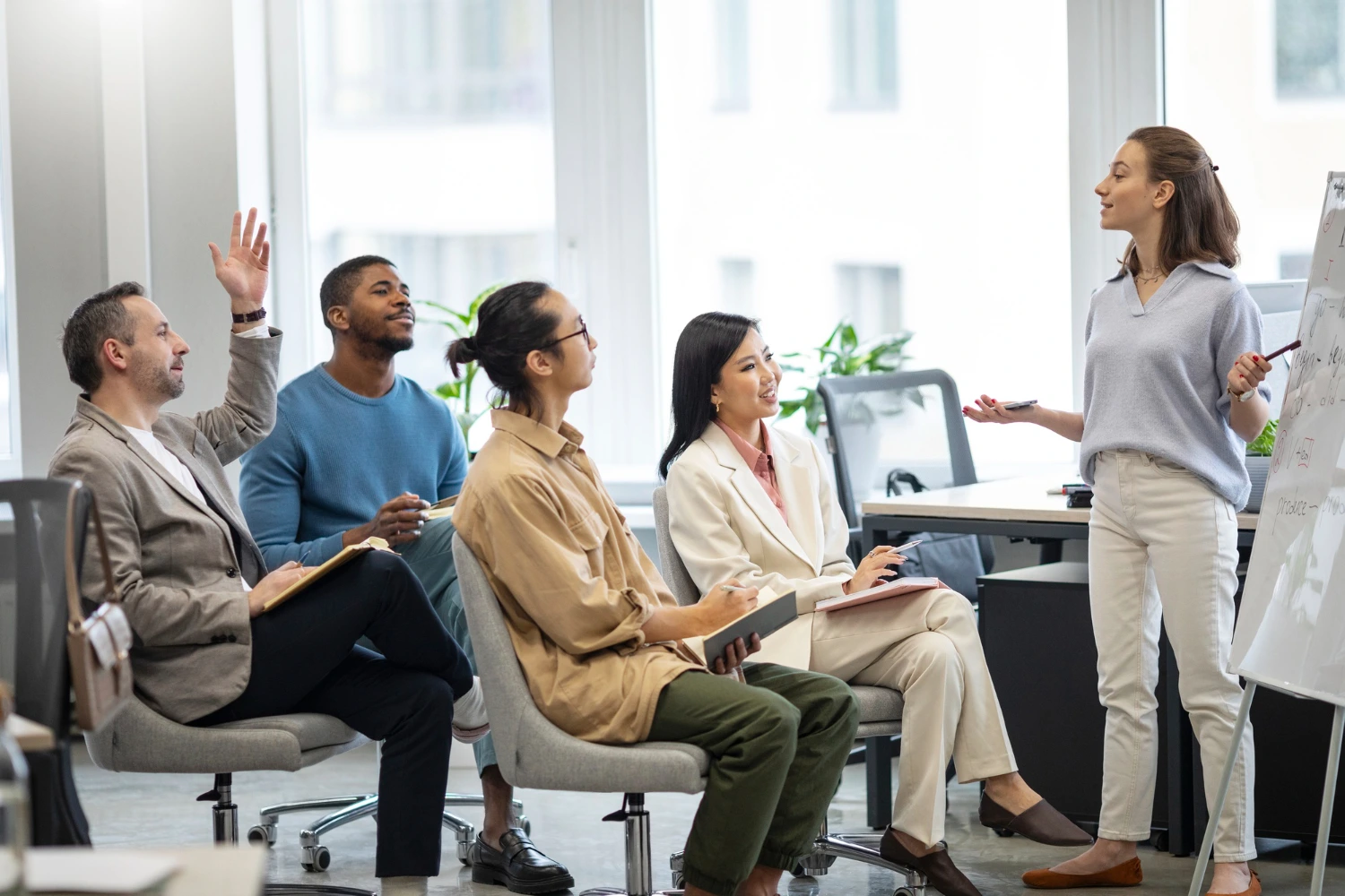 Uma equipe reunida em uma sala de reunião em treinamento sobre as atualizações da NR1. Uma mulher está apresentando informações em um quadro branco. Outros membros da equipe, sentados em cadeiras giratórias, prestam atenção e interagem, com um homem levantando a mão para fazer uma pergunta.