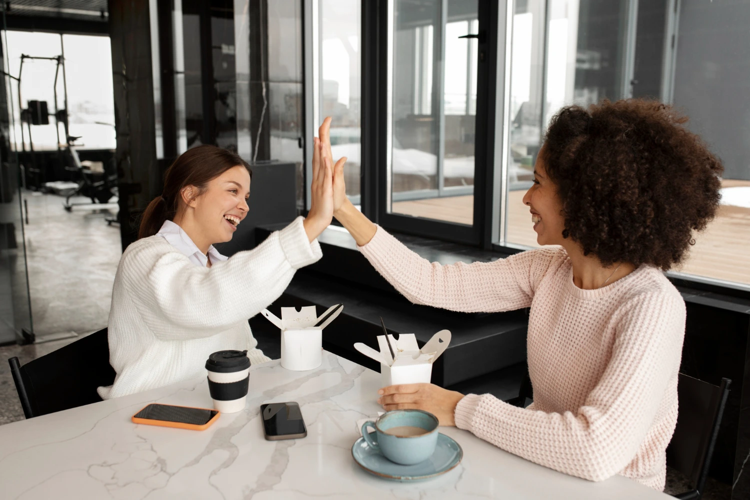 Duas mulheres estão sentadas em um café ou espaço de trabalho moderno, com grandes janelas permitindo muita luz natural. Elas dão as mãos no ar, expressando entusiasmo e celebração. Na mesa, há xícaras de café, recipientes de comida e outros itens pessoais, criando um ambiente colaborativo e positivo. A imagem ressalta a importância da interação positiva entre colegas, contribuindo para um ambiente saudável e produtivo, conforme NR-1.