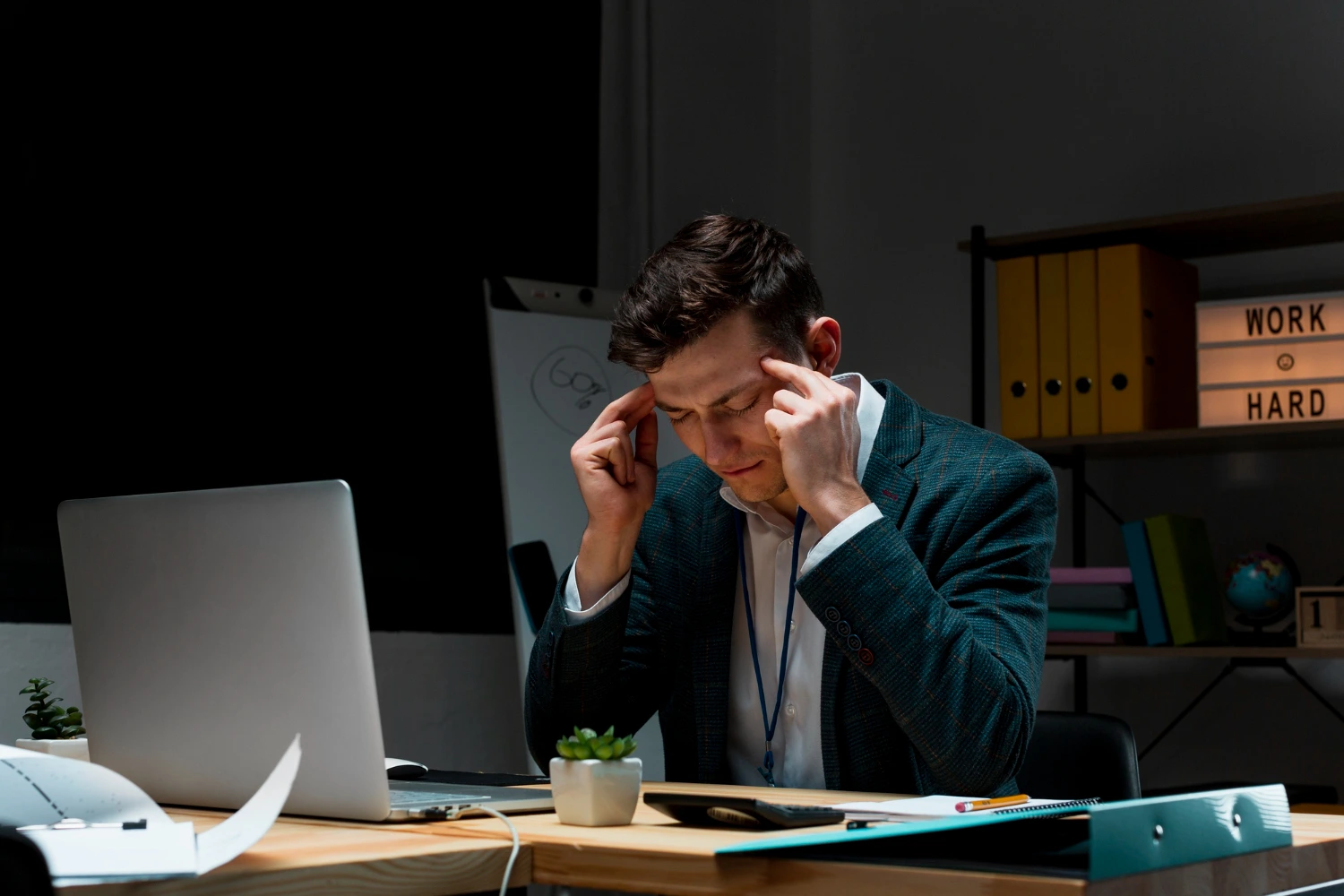Um homem vestindo um terno formal está sentado em frente a um laptop, com os dedos pressionando as têmporas, demonstrando sinais de cansaço ou estresse. O ambiente sugere uma carga excessiva de trabalho, destacando a importância de prevenir sobrecarga no ambiente laboral, conforme NR-1.