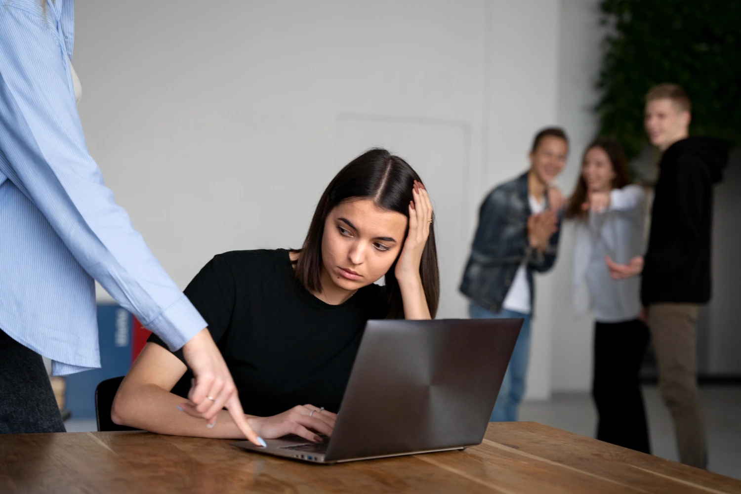 Funcionária sentada em frente ao notebook com expressão de desânimo enquanto colegas ao fundo riem e apontam – riscos psicossociais no trabalho.
