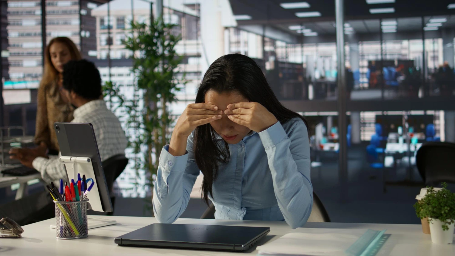 Mulher de cabeça baixa na mesa do escritório, com as mãos no rosto, expressando exaustão – riscos psicossociais no trabalho associados ao estresse contínuo.