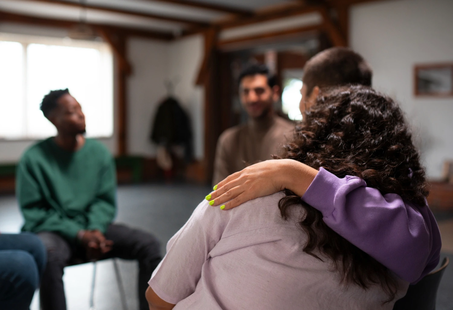 Uma mulher com cabelos cacheados e uma camiseta roxa abraça outra pessoa em um ambiente de sala, enquanto outras pessoas sorriem ao fundo. O cenário sugere apoio emocional e conexão social, destacando a importância da saúde mental no trabalho.