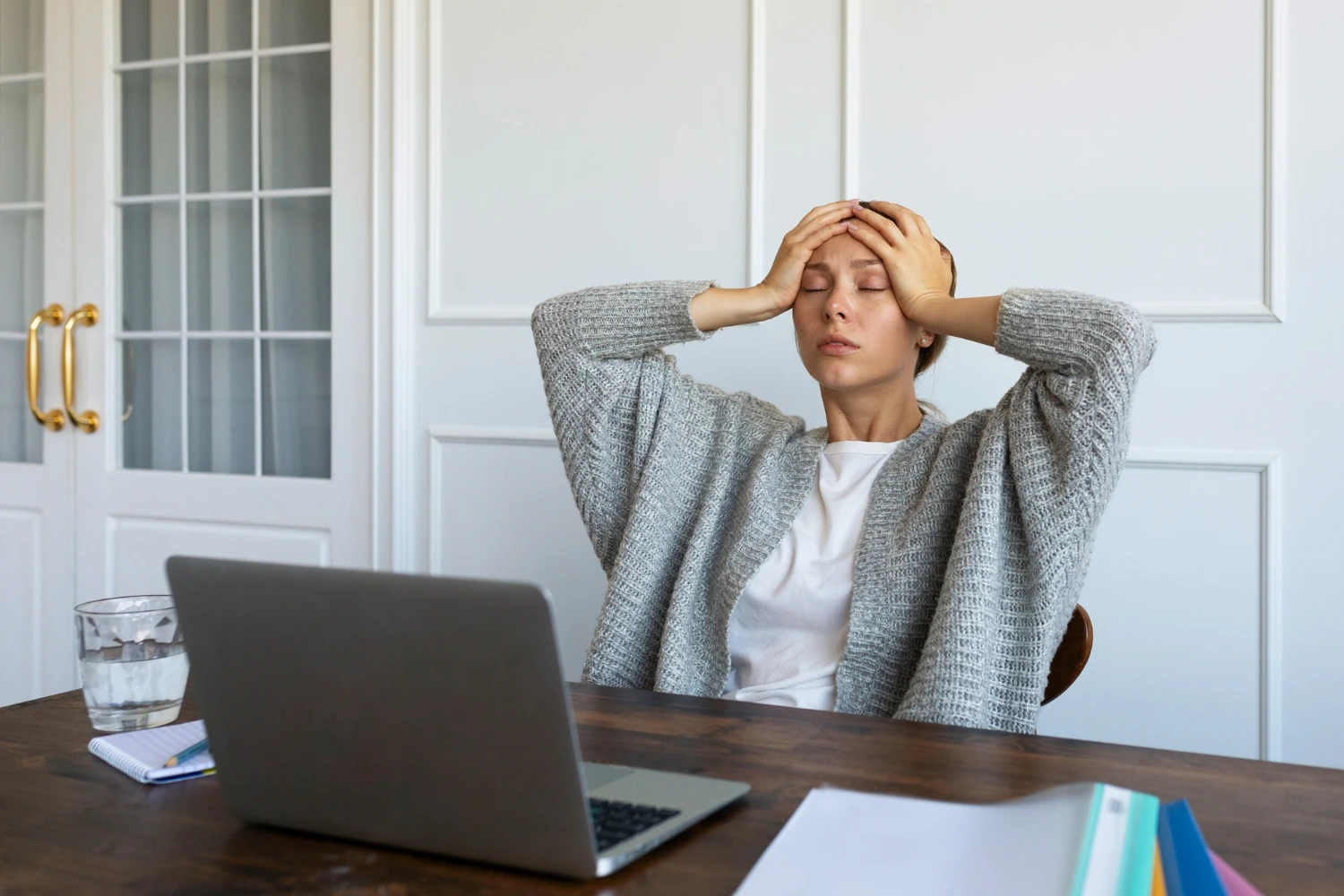 Uma mulher sentada à mesa de trabalho, segurando a cabeça com as mãos, demonstrando sinais de exaustão ou tensão. À sua frente, há um laptop e outros materiais de escritório, sugerindo que o estresse relacionado ao trabalho pode afetar diretamente a saúde mental.