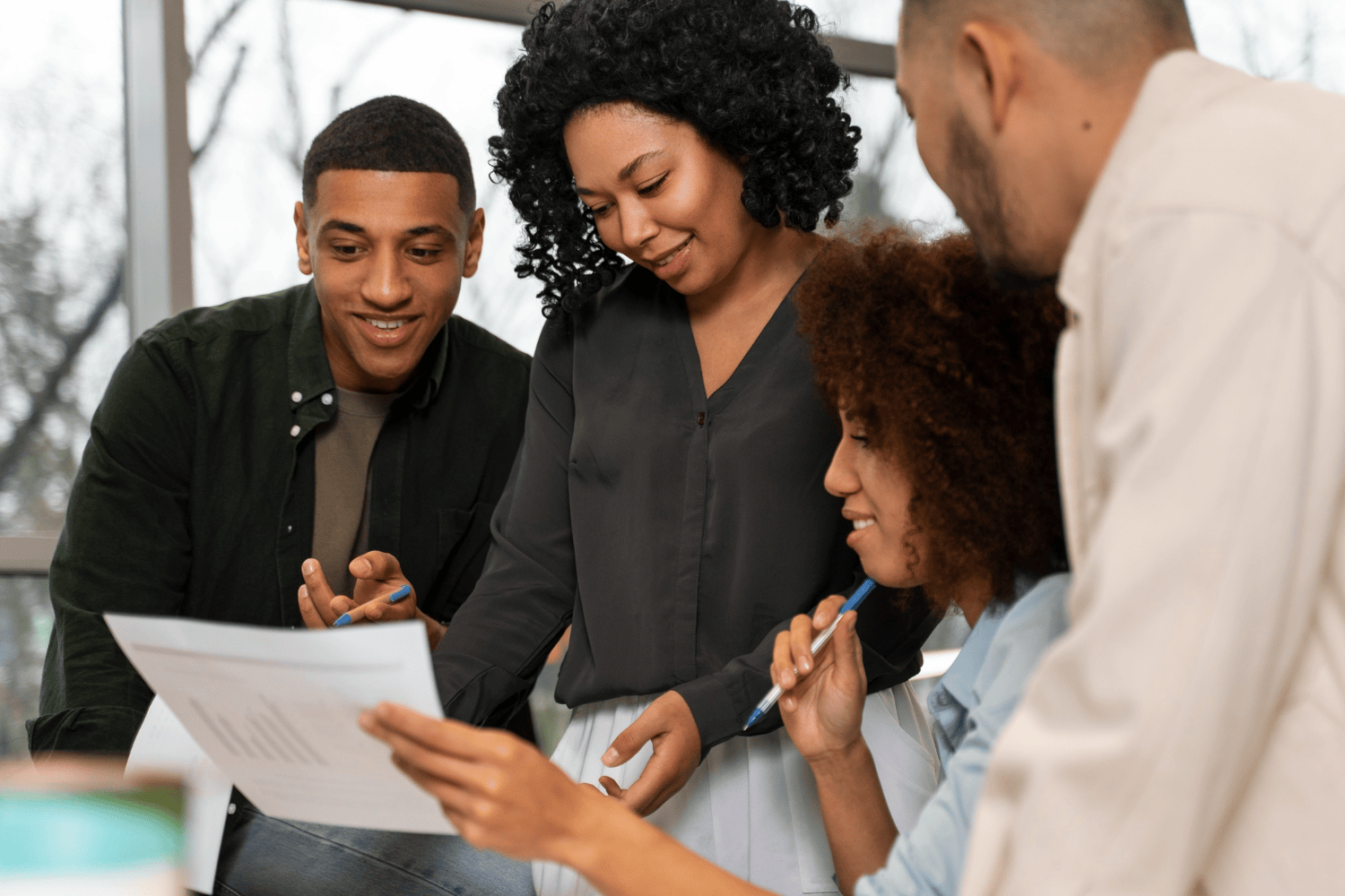 Grupo de quatro pessoas reunidas em ambiente corporativo, analisando documentos e trocando ideias de forma colaborativa. Uma mulher em pé lidera a conversa com atenção e escuta ativa, representando uma liderança de sucesso baseada no diálogo e no trabalho em equipe.