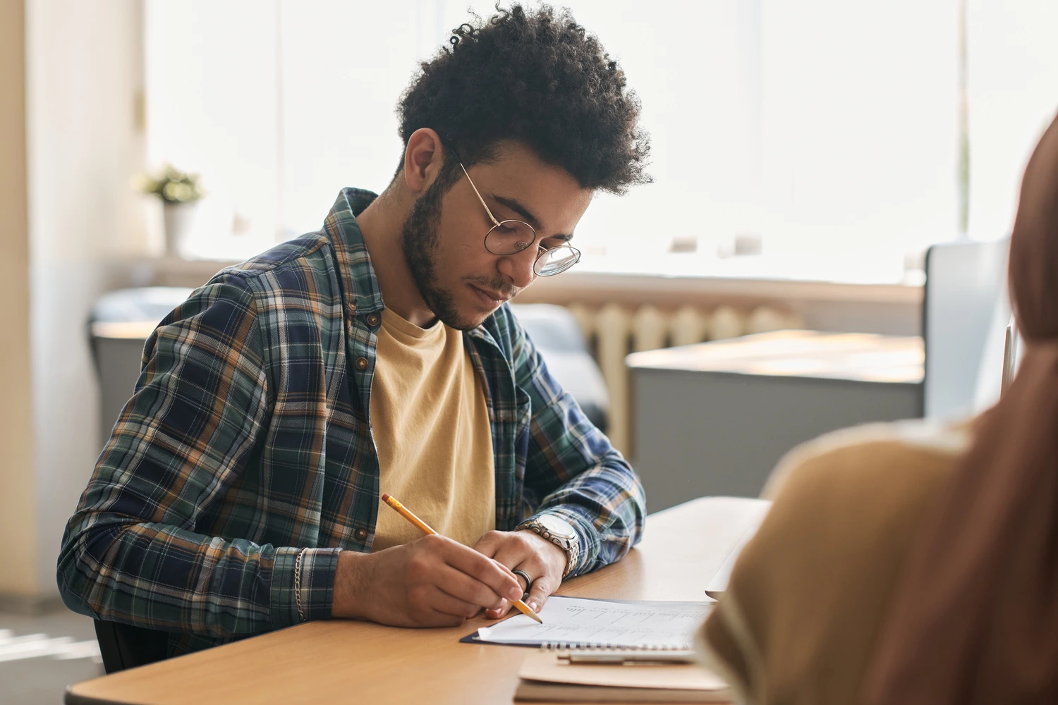 Um homem negro com cabelo encaracolado e óculos está sentado à mesa, concentrado em anotar algo em um caderno, concentrado em se preparar para Prova Nacional Docente. Ele veste uma camisa xadrez e usa um lápis para escrever.