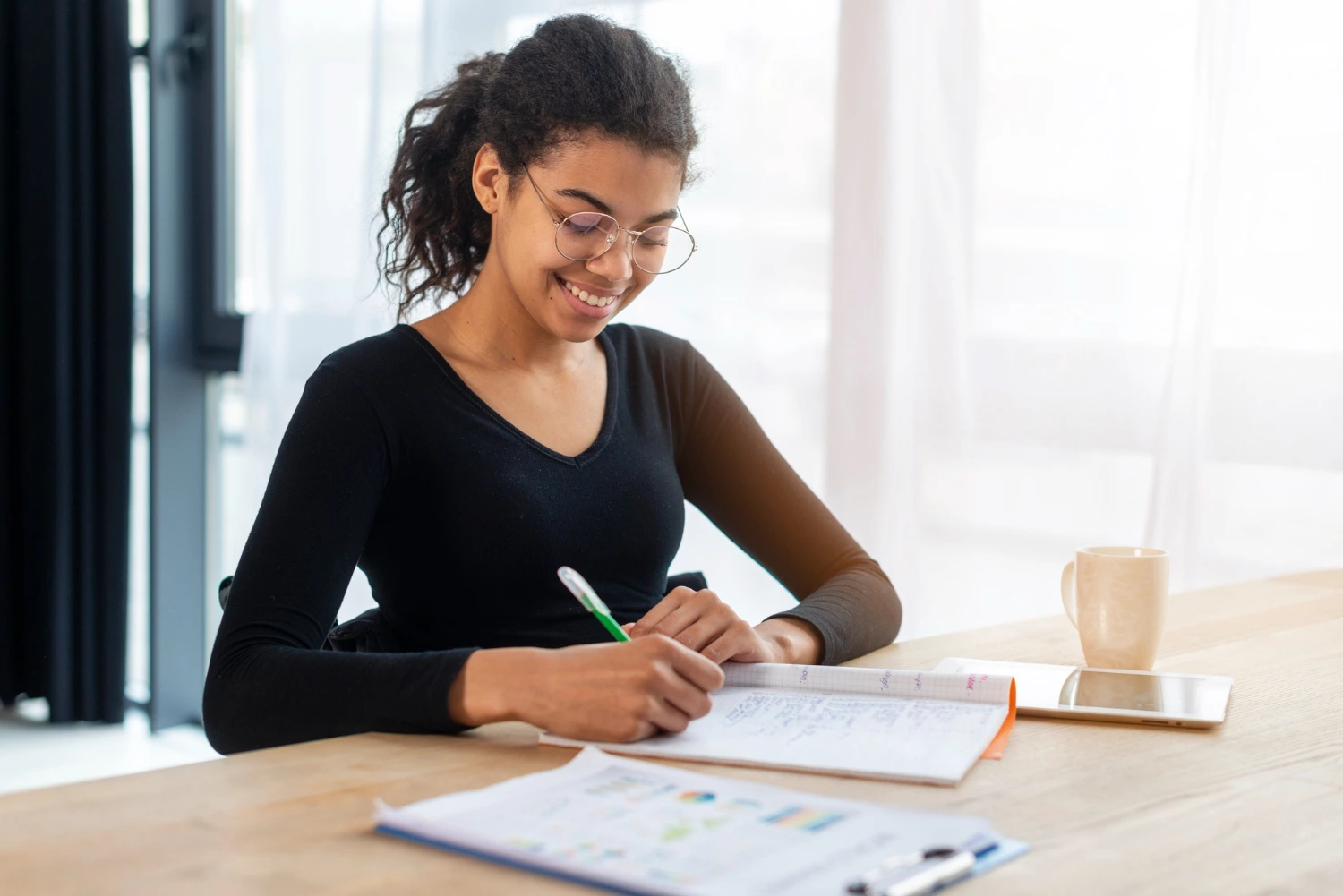 Uma jovem negra usando óculos e uma camiseta preta está sorrindo enquanto escreve em um caderno aberto sobre a mesa, ao estudar para a Prova Nacional Docente.