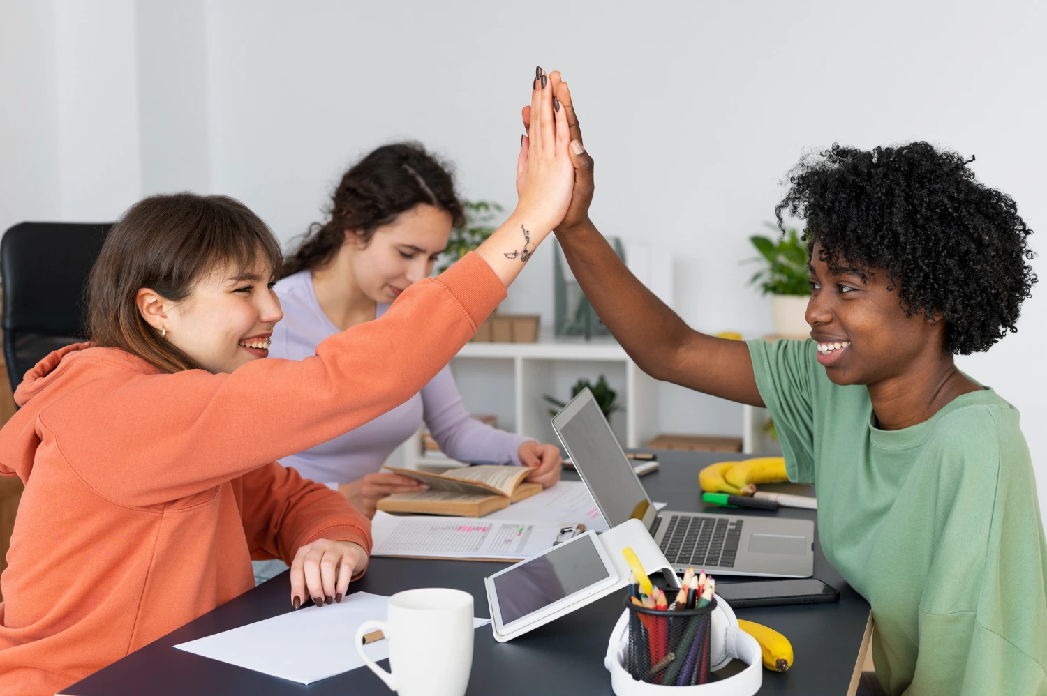 Duas mulheres comemoram em ambiente de trabalho com notebooks e livros, representando o empreendedorismo digital e a flexibilidade no trabalho.