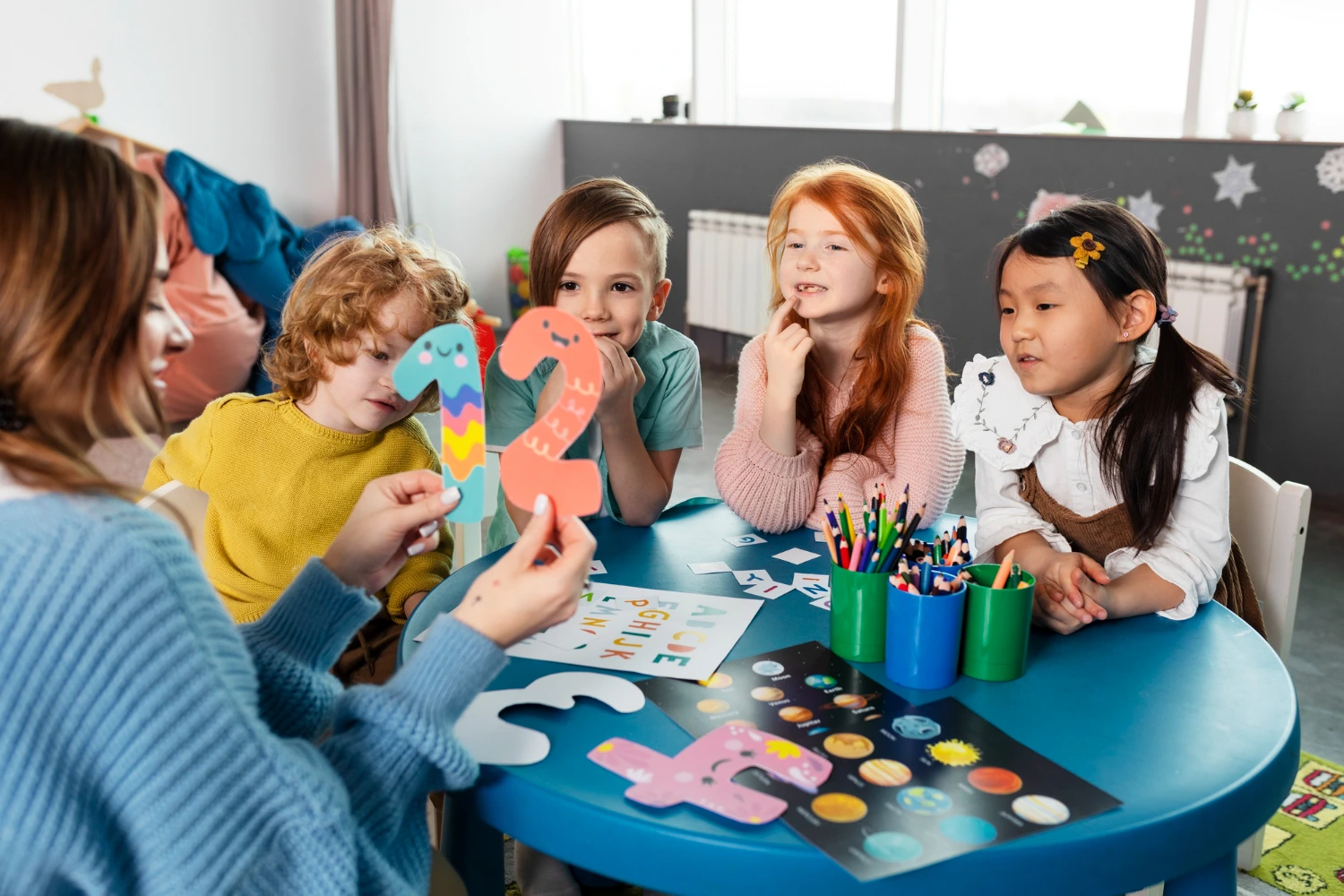 Crianças observam números coloridos em atividade educativa com professora, em sala decorada com materiais pedagógicos sobre planetas e alfabetização.