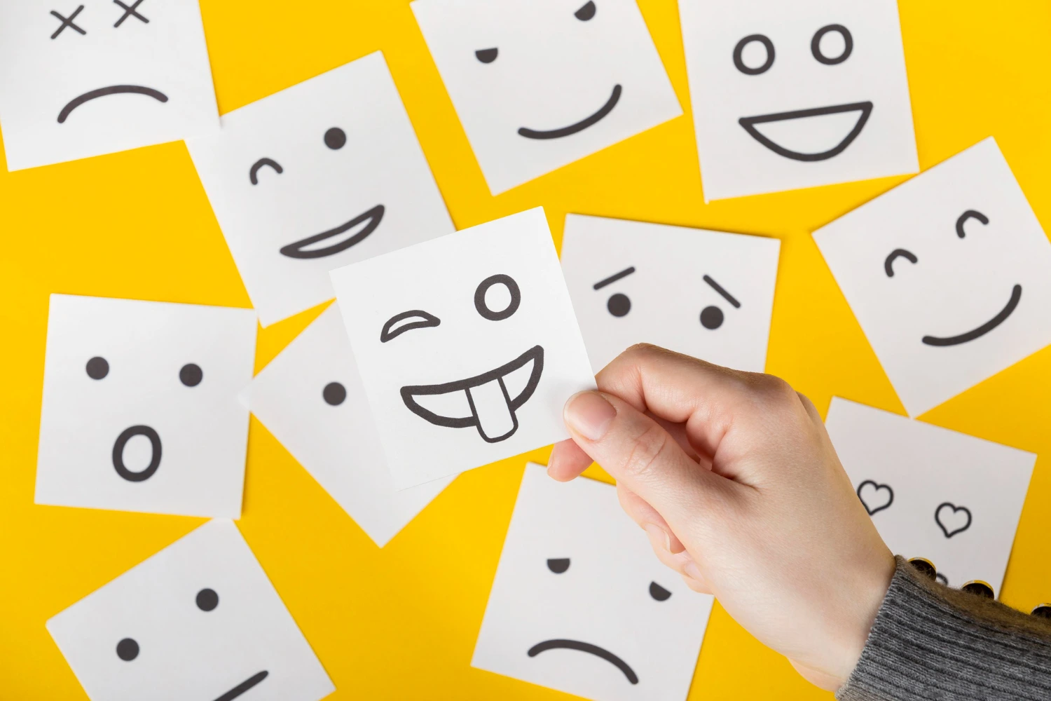 Mão segura papel com emoji divertido sobre fundo amarelo repleto de expressões faciais. Sugere características de um líder com inteligência emocional e capacidade de lidar com sentimentos.