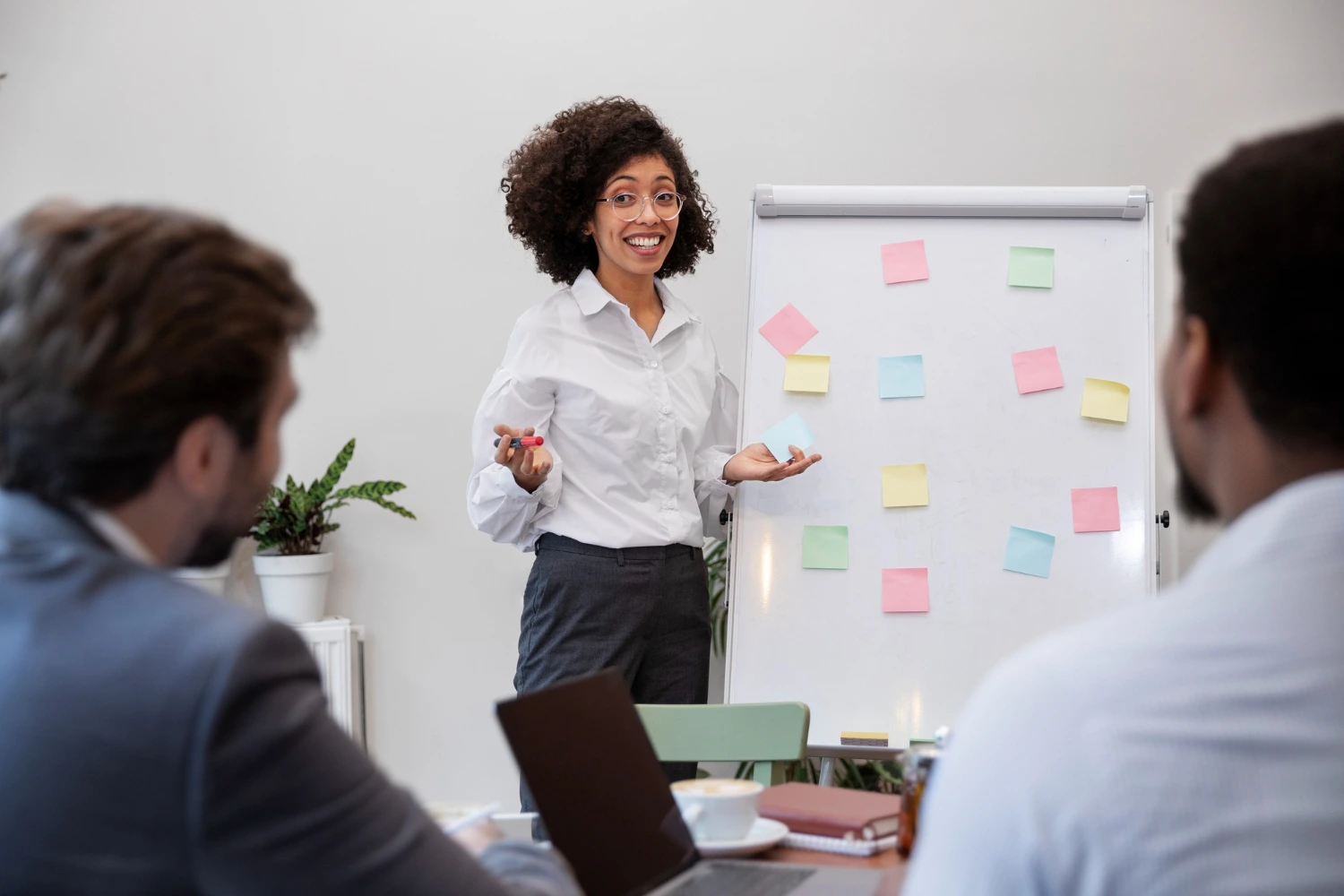 Mulher apresenta ideias em quadro branco com post-its coloridos, diante de colegas atentos. Destaca características de um líder que organiza, planeja e conduz reuniões de forma clara.