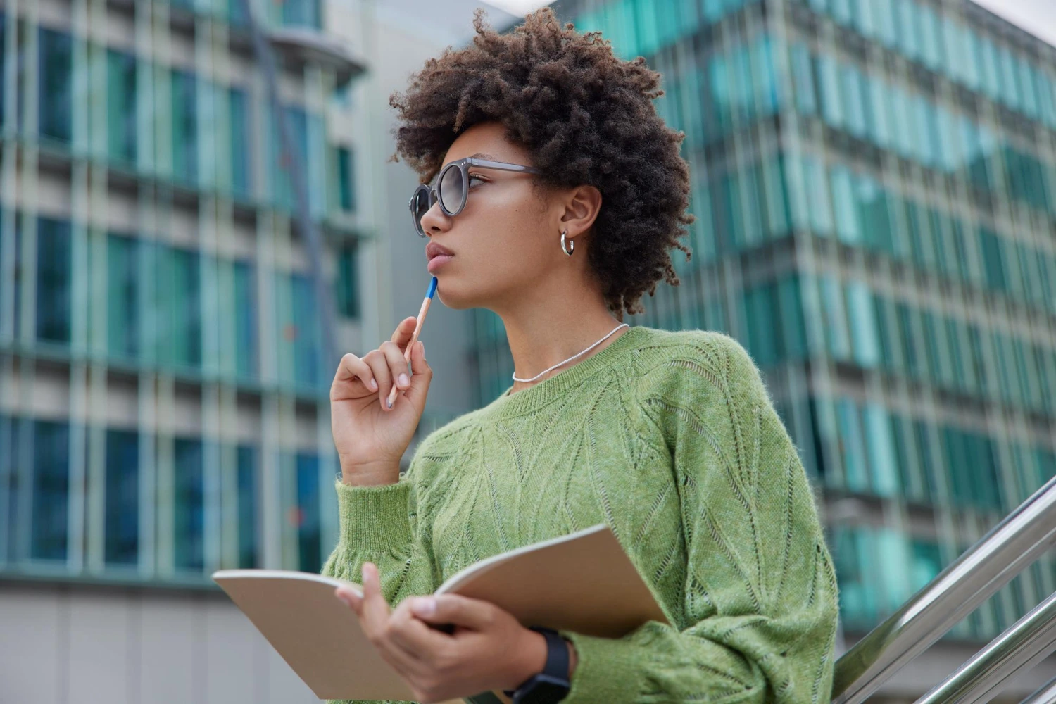 Uma jovem com cabelo cacheado volumoso e óculos escuros está em pé em frente a um prédio de vidro moderno. Ela veste um suéter verde e segura um caderno aberto em uma das mãos. Com a outra mão, leva uma caneta azul à boca em gesto pensativo, olhando para frente como se refletisse sobre sua carreira em Y.