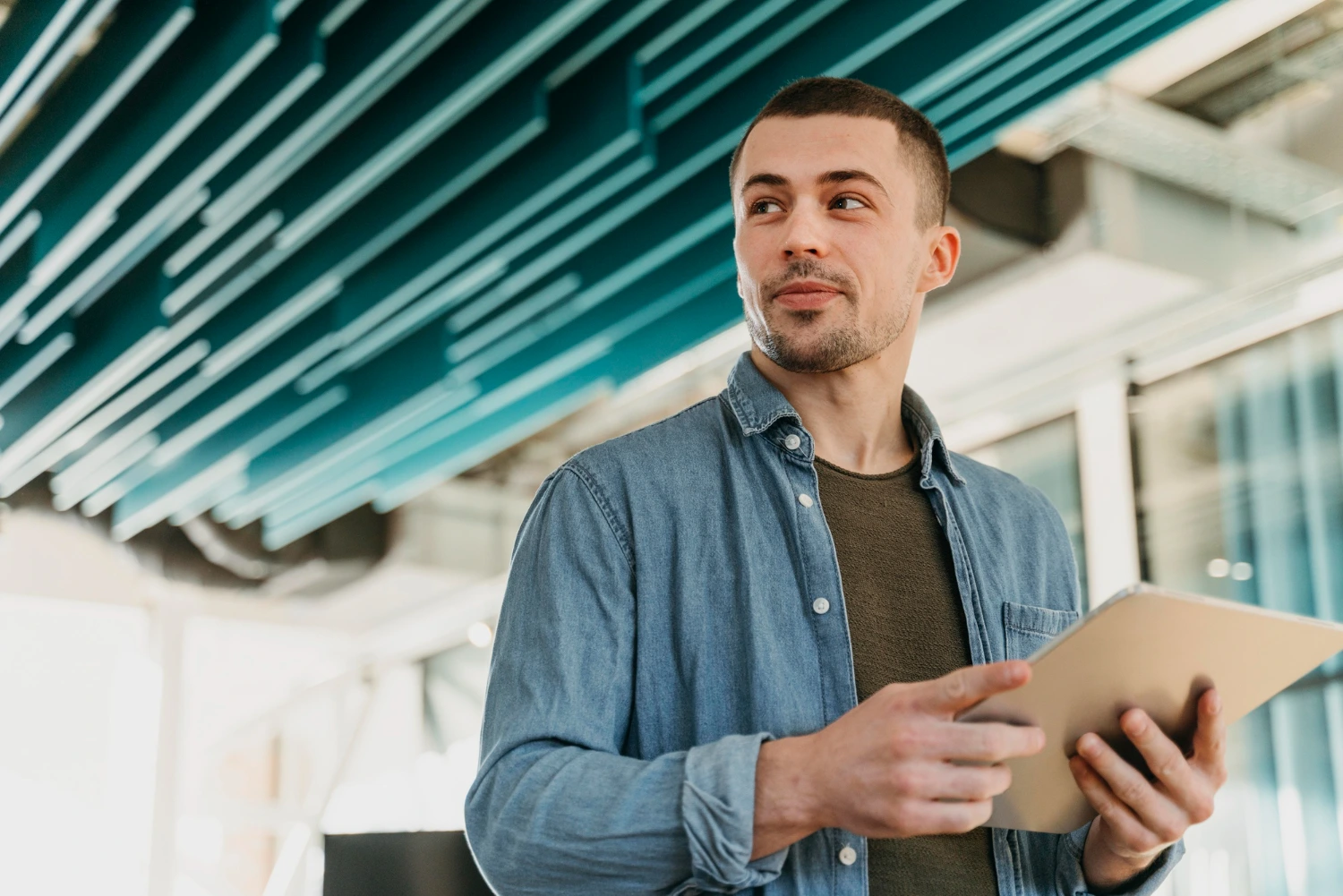 Um homem jovem, vestido com camisa jeans, segura um tablet enquanto observa algo ao redor em um ambiente moderno. A imagem sugere inovação e mobilidade no trabalho, destacando como o pacote office também pode ser acessado em dispositivos móveis, facilitando a produtividade em qualquer lugar.