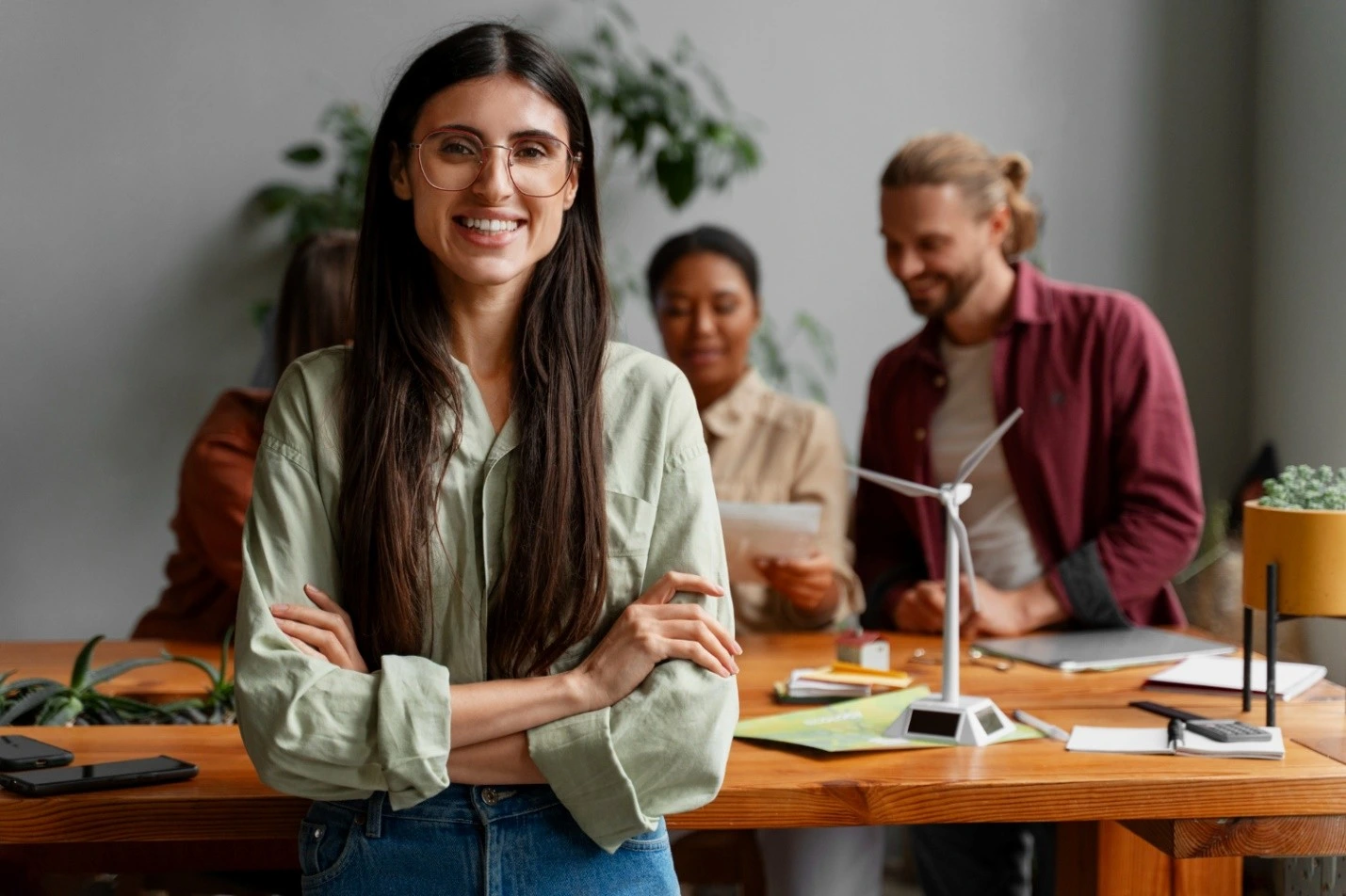 Uma jovem de óculos e cabelo comprido sorri em primeiro plano com os braços cruzados, em um ambiente de trabalho colaborativo. Ao fundo, três colegas conversam animadamente ao redor de uma mesa com maquete de turbina eólica e materiais de estudo. A imagem remete a inovação, energia limpa e trabalho em equipe, elementos centrais para quem busca se especializar por meio de cursos de ESG.