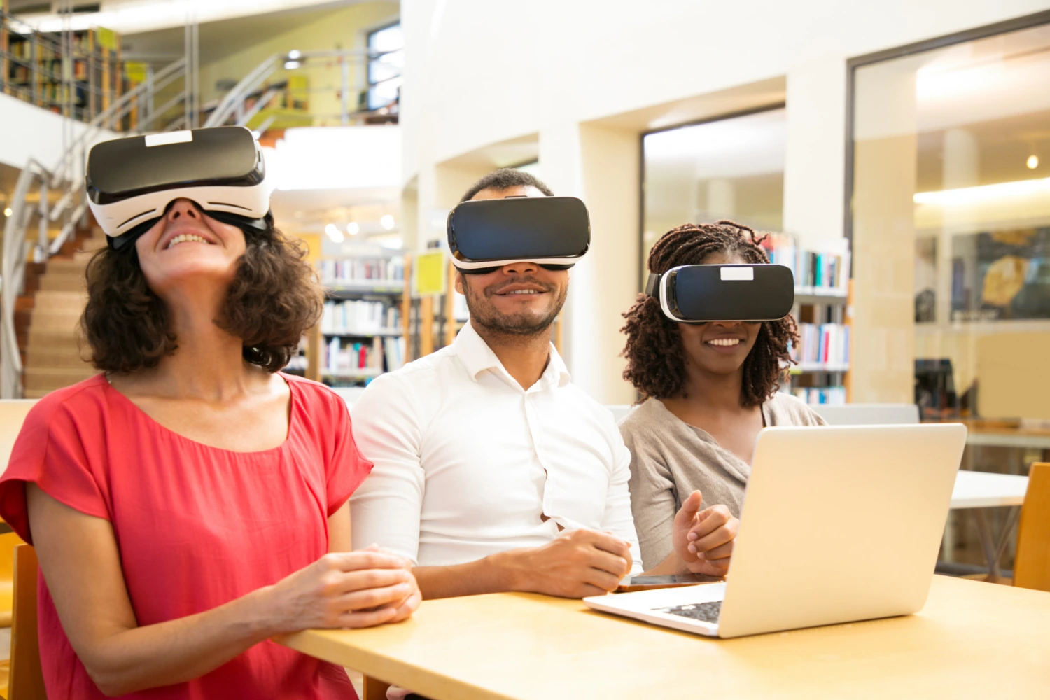 Três pessoas estão sentadas em uma mesa utilizando óculos de realidade virtual, sorrindo e interagindo com a experiência digital. Um notebook está aberto à frente, sugerindo integração entre tecnologia e aprendizado. A cena representa como recursos imersivos fazem parte do futuro da educação, trazendo inovação e engajamento para alunos de todas as idades.