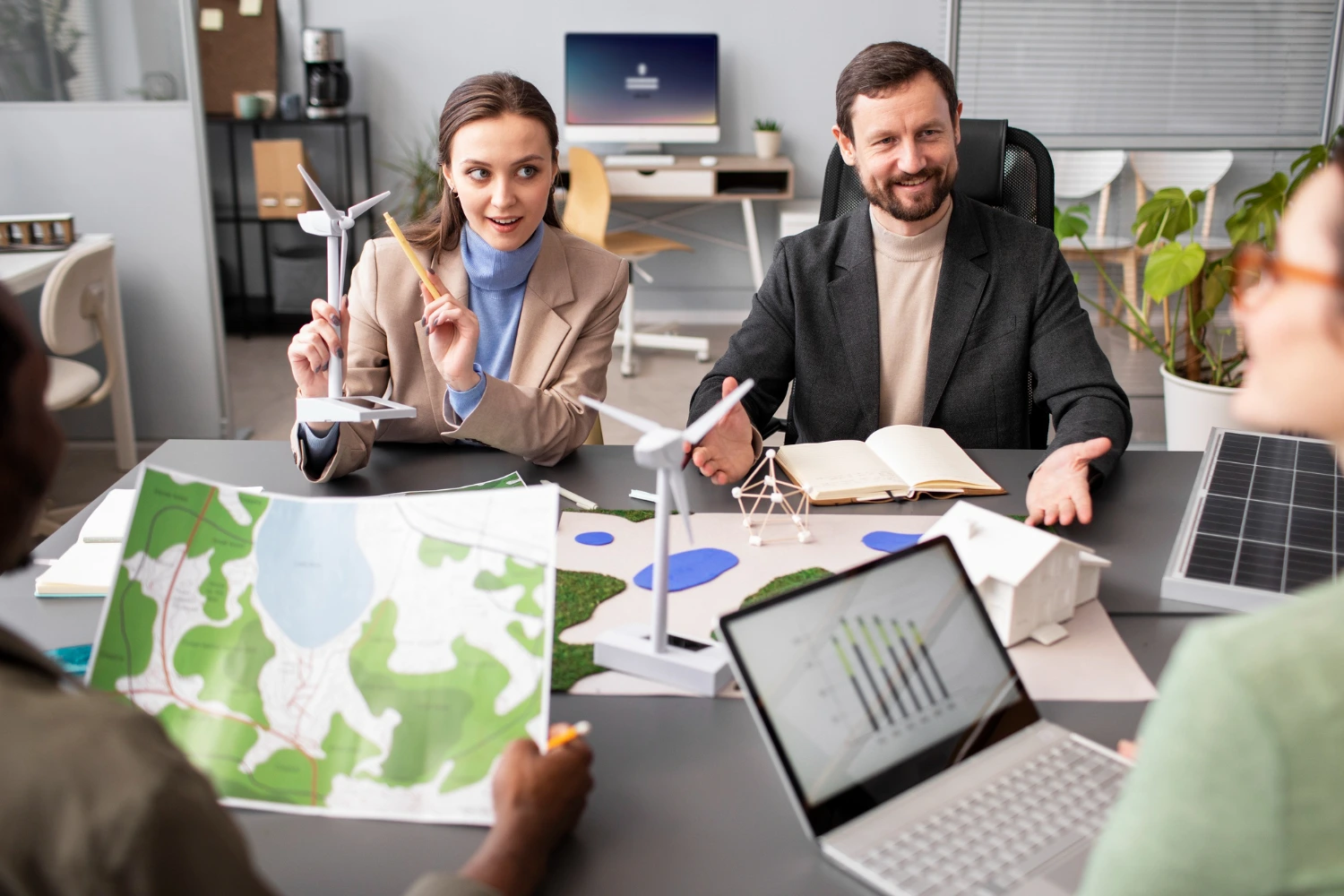 Quatro pessoas participam de uma reunião em um escritório moderno, analisando mapas, maquetes de turbinas eólicas e painéis solares. A interação mostra o trabalho em equipe na busca por soluções ambientais inovadoras, reforçando a importância dos profissionais especializados em ESG na criação de projetos sustentáveis e de impacto positivo.