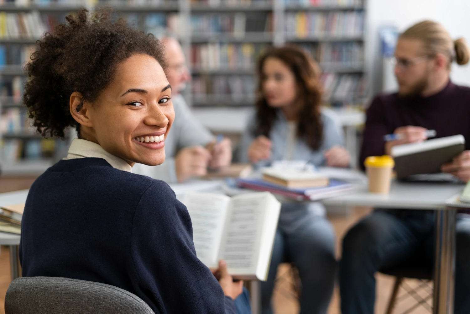 Uma estudante sorri para a câmera enquanto segura um livro aberto, em meio a um grupo de colegas que conversam e estudam em uma biblioteca. O ambiente acadêmico reforça o clima de preparação e colaboração, destacando como os concursos públicos na educação podem transformar trajetórias profissionais e abrir novas oportunidades de carreira.